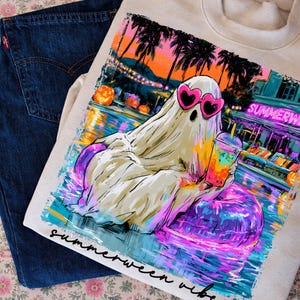 May include: A cream-coloured sweatshirt featuring a graphic of a ghost wearing pink heart-shaped sunglasses, holding a colourful drink while floating in a pool on a purple inner tube, with palm trees and the text "SUMMERWEEN" and "summerween vibe".