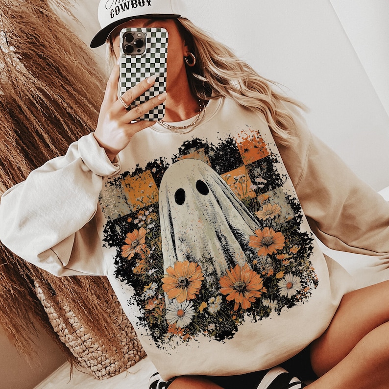 Puede incluir: Sudadera beige con un dise&ntilde;o de fantasma y flores naranjas y blancas. El fantasma tiene dos ojos negros y est&aacute; rodeado de un arreglo floral. La sudadera tiene mangas largas y cuello redondo.