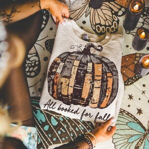 Pu&ograve; includere: Felpa beige con un design a forma di zucca fatta di libri marroni, arancioni e grigi. Il testo "All booked for fall" &egrave; stampato sotto. La felpa &egrave; piegata su una superficie a fantasia. L'immagine include candele accese.
