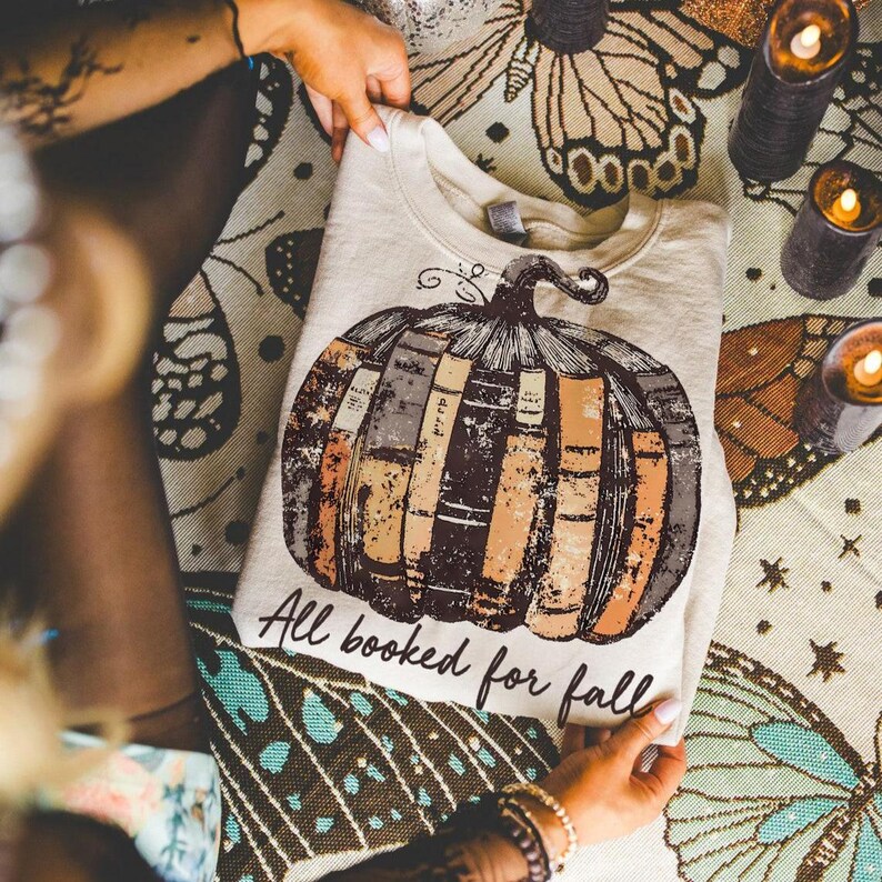 Pu&ograve; includere: Felpa beige con un design a forma di zucca fatta di libri marroni, arancioni e grigi. Il testo "All booked for fall" &egrave; stampato sotto. La felpa &egrave; piegata su una superficie a fantasia. L'immagine include candele accese.