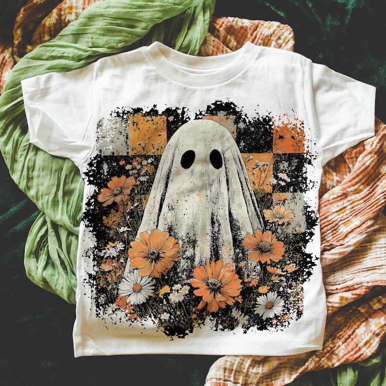 Puede incluir: Camiseta blanca con un dise&ntilde;o de fantasma con agujeros negros para los ojos, rodeado de flores naranjas y blancas. El fantasma est&aacute; sobre un fondo a cuadros de cuadrados naranjas, negros y grises. La camiseta tiene mangas cortas.