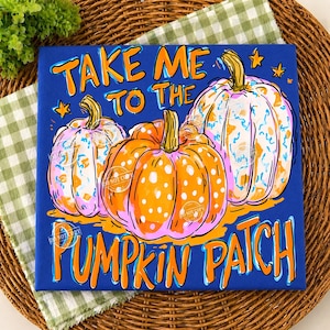 May include: A square sign with a blue background featuring three pumpkins in orange, pink, and white with blue and orange speckles. The text reads "TAKE ME TO THE PUMPKIN PATCH" in orange lettering. Small orange stars are scattered around the pumpkins.