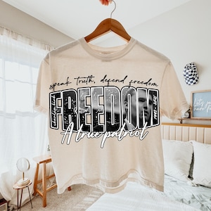 May include: A cream-coloured t-shirt with the word "FREEDOM" in a bold, black and white design. The shirt features a black and white portrait and the text "speak truth, defend freedom" and "A True Patriot".