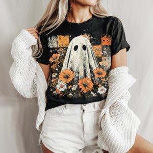 Puede incluir: Camiseta negra con un dise&ntilde;o de fantasma rodeado de flores naranjas y blancas. El fantasma tiene dos ojos negros. La camiseta tiene un aspecto vintage y desgastado. Un c&aacute;rdigan blanco est&aacute; sobre los hombros.
