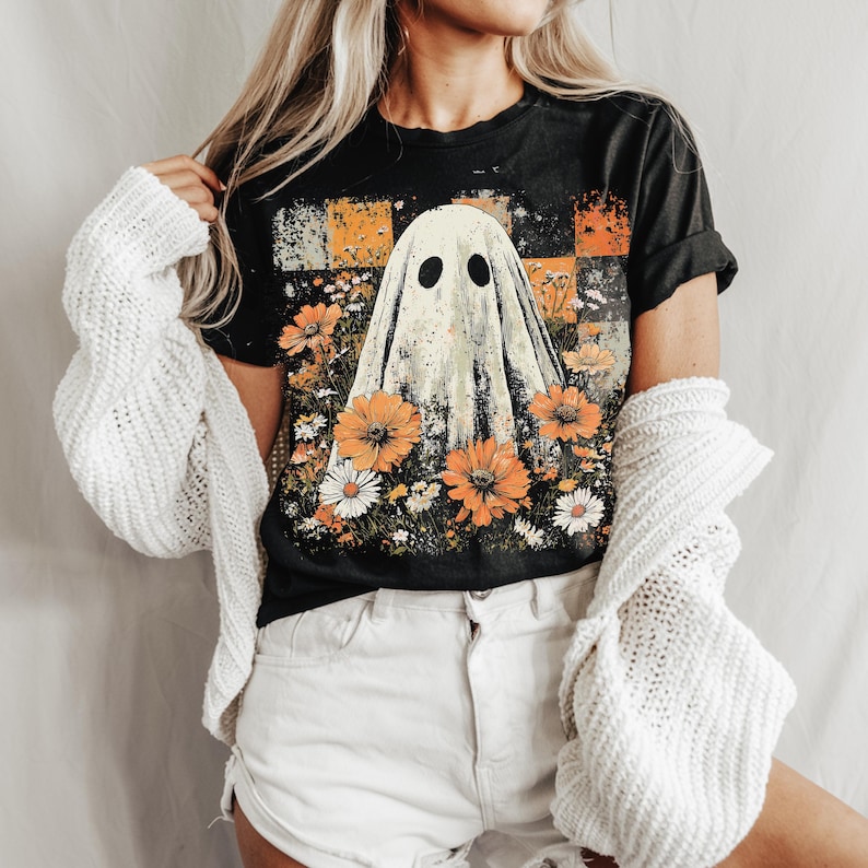 Puede incluir: Camiseta negra con un dise&ntilde;o de fantasma rodeado de flores naranjas y blancas. El fantasma tiene dos ojos negros. La camiseta tiene un aspecto vintage y desgastado. Un c&aacute;rdigan blanco est&aacute; sobre los hombros.