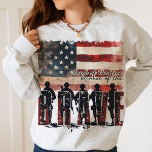 May include: A gray sweatshirt with a distressed American flag design and the text "land of the free because of the brave". The flag is red, white, and blue with five rows of stars. Five silhouettes of soldiers are standing in front of the flag, forming the word "brave".