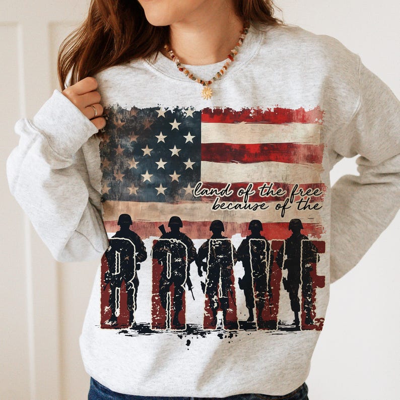 May include: A gray sweatshirt with a distressed American flag design and the text "land of the free because of the brave". The flag is red, white, and blue with five rows of stars. Five silhouettes of soldiers are standing in front of the flag, forming the word "brave".