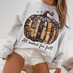Pu&ograve; includere: Felpa grigio chiaro con un design a zucca fatto di libri nei toni del marrone, arancione e giallo. Il testo "All booked for fall" &egrave; stampato sotto. Un capo d'abbigliamento a tema autunnale.