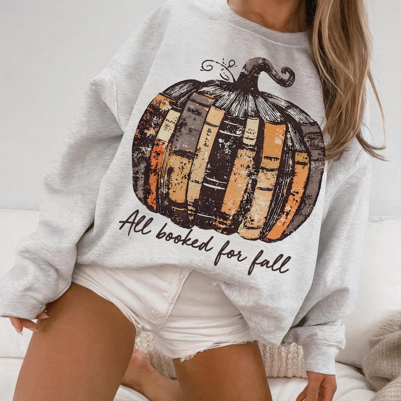 Pu&ograve; includere: Felpa grigio chiaro con un design a zucca fatto di libri nei toni del marrone, arancione e giallo. Il testo "All booked for fall" &egrave; stampato sotto. Un capo d'abbigliamento a tema autunnale.