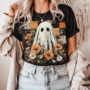 Puede incluir: Camiseta negra con un dise&ntilde;o de fantasma rodeado de flores naranjas y blancas. El fantasma tiene dos ojos ovalados negros. El fondo presenta un patr&oacute;n de cuadros en naranja y amarillo, con un estilo casual.