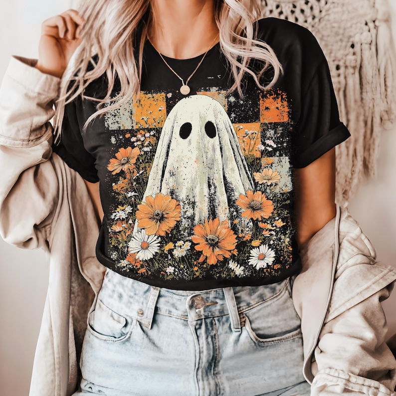 Puede incluir: Camiseta negra con un dise&ntilde;o de fantasma rodeado de flores naranjas y blancas. El fantasma tiene dos ojos ovalados negros. El fondo presenta un patr&oacute;n de cuadros en naranja y amarillo, con un estilo casual.