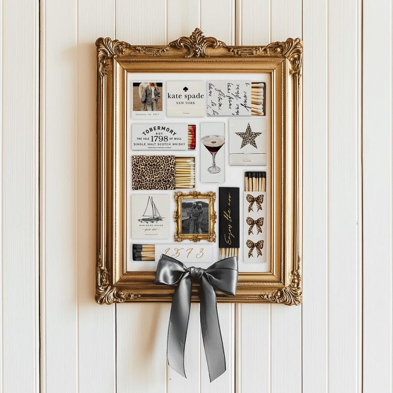 May include: A gold framed picture with a white background and various matchboxes inside. The matchboxes have different designs, including a leopard print, a sailboat, a martini glass, and a star. The frame is decorated with a gray bow.
