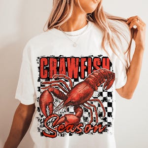 May include: White t-shirt with a red crawfish graphic and the text "Crawfish Season" in black and white checkered background.