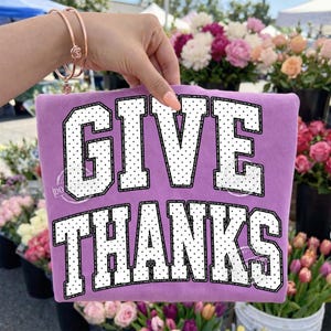 Op de afbeelding: Een lavendelkleurig stoffen item met de tekst GIVE THANKS in grote, witte blokletters met een zwart stippenpatroon en een dikke zwarte omlijning. De tekst is in twee rijen gerangschikt, met GIVE bovenaan en THANKS daaronder.