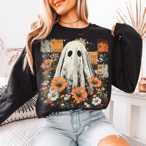 Puede incluir: Sudadera negra de manga larga con un dise&ntilde;o de fantasma y un arreglo floral. El fantasma tiene dos agujeros negros para los ojos y est&aacute; rodeado de flores naranjas y blancas. El fondo tiene un patr&oacute;n de cuadros en naranja, negro y blanco.