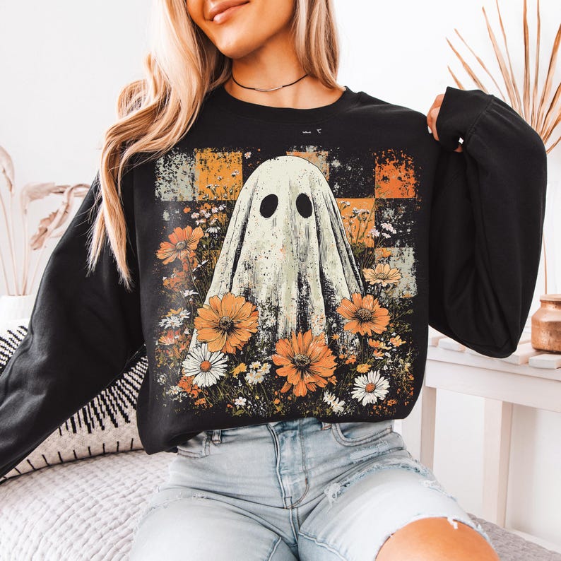 Puede incluir: Sudadera negra de manga larga con un dise&ntilde;o de fantasma y un arreglo floral. El fantasma tiene dos agujeros negros para los ojos y est&aacute; rodeado de flores naranjas y blancas. El fondo tiene un patr&oacute;n de cuadros en naranja, negro y blanco.
