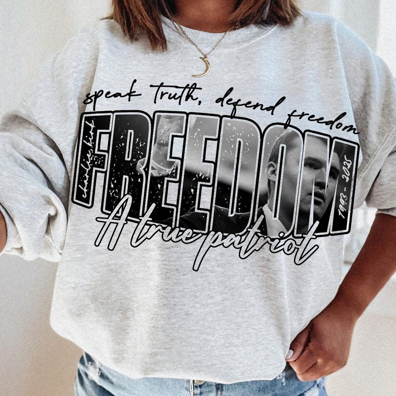 May include: Pale grey sweatshirt with the word "FREEDOM" in a distressed black and white design, featuring a portrait. The text "speak truth, defend freedom" and "A true patriot" are also displayed.