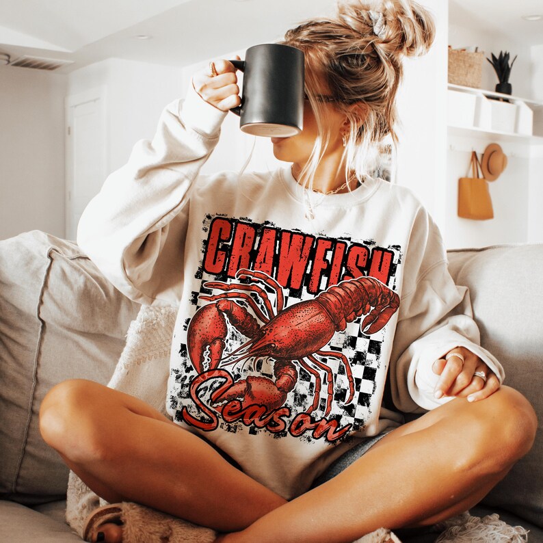 May include: A white sweatshirt with a red crawfish graphic and the text "Crawfish Season" on a black and white checkered background.