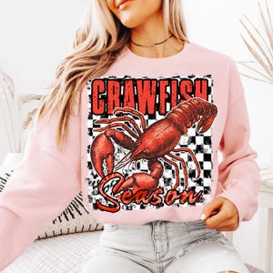 May include: Pink sweatshirt with a red crawfish graphic and the text "Crawfish Season" on a black and white checkered background.