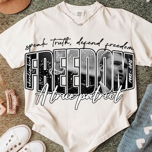 May include: Off-white t-shirt featuring the word "FREEDOM" in large, bold, black letters, with a black and white image of a person inside the letters. The text "speak truth, defend freedom" is above the word "FREEDOM". The text "A true patriot" is below the word "FREEDOM".