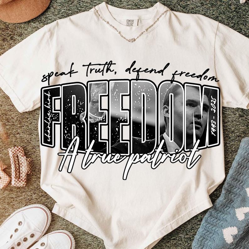 May include: Off-white t-shirt featuring the word "FREEDOM" in large, bold, black letters, with a black and white image of a person inside the letters. The text "speak truth, defend freedom" is above the word "FREEDOM". The text "A true patriot" is below the word "FREEDOM".