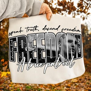 May include: A cream-coloured tote bag with the word "FREEDOM" in large, bold, black and white letters. The bag features a black and white portrait of a man and the text "Speak truth, defend freedom" and "A true patriot".