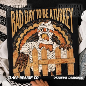 May include: A black t-shirt featuring a graphic of a turkey wrapped in white bandages like a mummy, standing behind a wooden fence. The text above the turkey reads BAD DAY TO BE A TURKEY in large, bold, gold-coloured, distressed-style lettering.