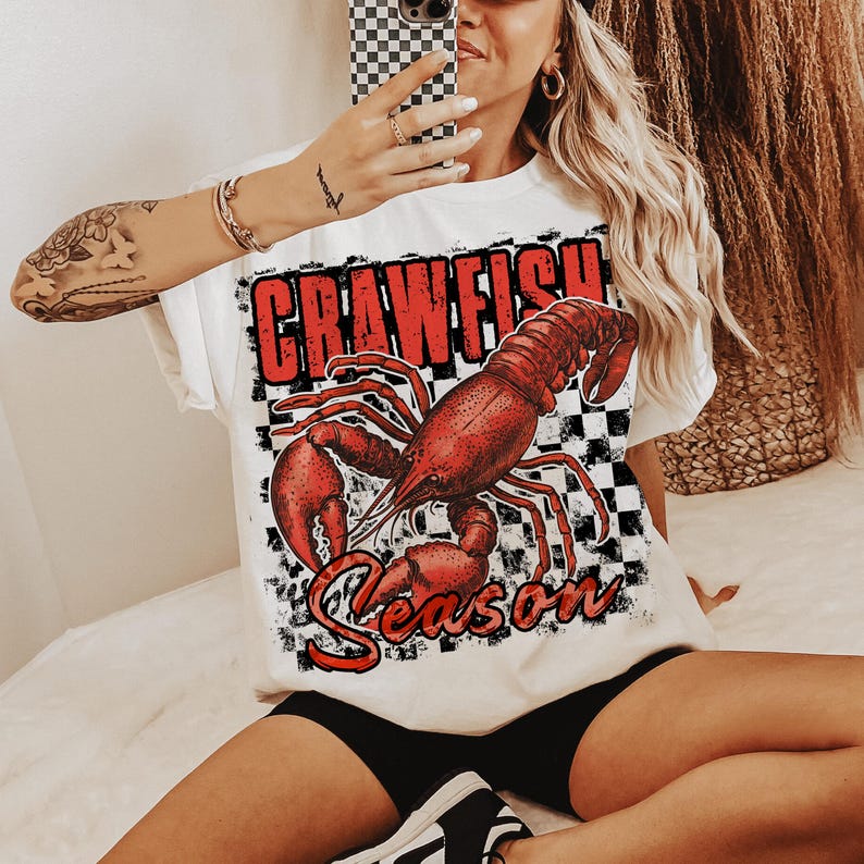 May include: White t-shirt with a red crawfish graphic and the text "Crawfish Season" in black. The crawfish is on a black and white checkered background.