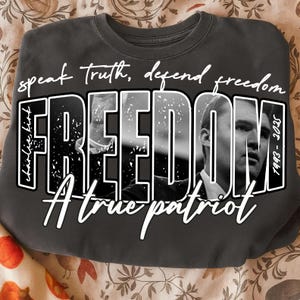 May include: Dark grey sweatshirt featuring the word "FREEDOM" in large, bold, white letters. The word contains a black and white image of a person. The text "speak truth, defend freedom" and "A true patriot" are also on the sweatshirt.