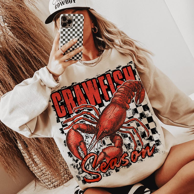 May include: A beige sweatshirt with a red crawfish graphic and the text "Crawfish Season" in black. The crawfish is on a black and white checkered background.