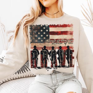 May include: A beige sweatshirt with a distressed American flag design and the text "land of the free because of the BRAVE".