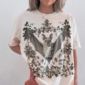 May include: Cream-colored t-shirt featuring a detailed illustration of a bat with outstretched wings, surrounded by floral and ornamental elements. The bat is depicted in grayscale, with the floral elements adding contrast. The shirt has short sleeves.