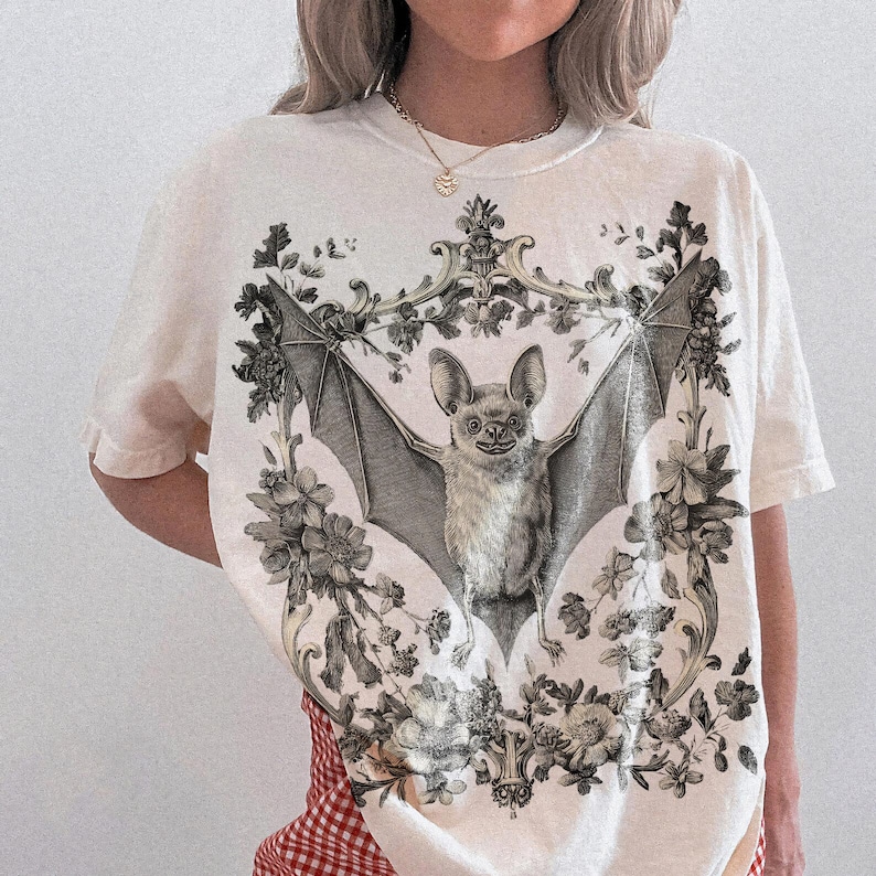 May include: Cream-colored t-shirt featuring a detailed illustration of a bat with outstretched wings, surrounded by floral and ornamental elements. The bat is depicted in grayscale, with the floral elements adding contrast. The shirt has short sleeves.