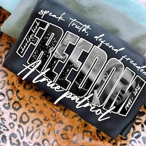 May include: A dark grey t-shirt with the word "FREEDOM" in a bold, distressed font, featuring a black and white portrait. The shirt also includes the phrases "speak truth, defend freedom" and "A true patriot."
