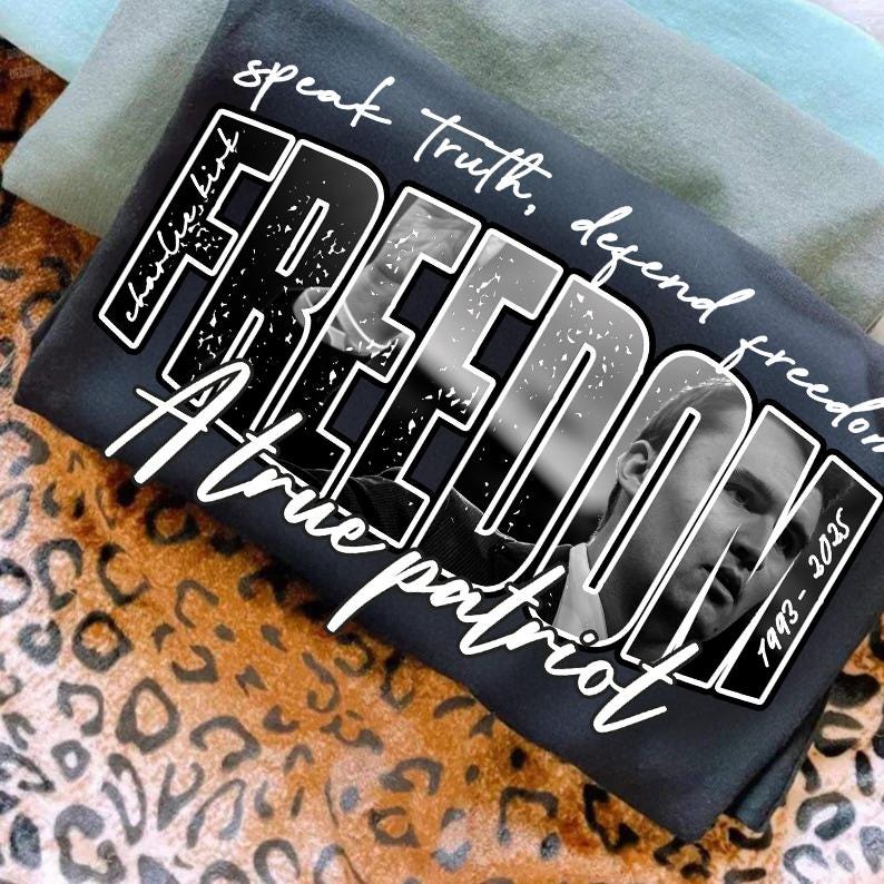 May include: A dark grey t-shirt with the word "FREEDOM" in a bold, distressed font, featuring a black and white portrait. The shirt also includes the phrases "speak truth, defend freedom" and "A true patriot."