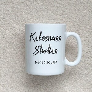 May include: A white ceramic mug with a curved handle. The mug features the text "Kokosnuss Studios" in a script font, with "MOCKUP" below. The mug is set against a neutral, textured background.