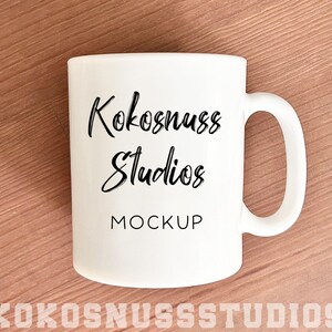 May include: A white ceramic mug with a curved handle. The mug features the text "Kokosnuss Studios MOCKUP" in a black, handwritten-style font. The mug is set against a light brown wooden surface, with the text "KOKOSNUSSSTUDIOS" at the bottom.