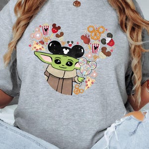 May include: A grey t-shirt with a graphic of Baby Yoda wearing Mickey Mouse ears and holding a lollipop. The design incorporates Mickey Mouse-themed treats such as popcorn, pretzels, and pizza, arranged in a heart shape.