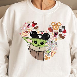 May include: White crew neck sweatshirt with a graphic of Grogu, or Baby Yoda, holding a lollipop. The design incorporates a Mickey Mouse head made of snacks. The sweatshirt is a casual style.