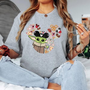 May include: A grey t-shirt with a graphic of Baby Yoda wearing Mickey Mouse ears and holding a lollipop. The design includes Mickey Mouse-themed treats like popcorn, pretzels, and pizza, arranged in a heart shape.