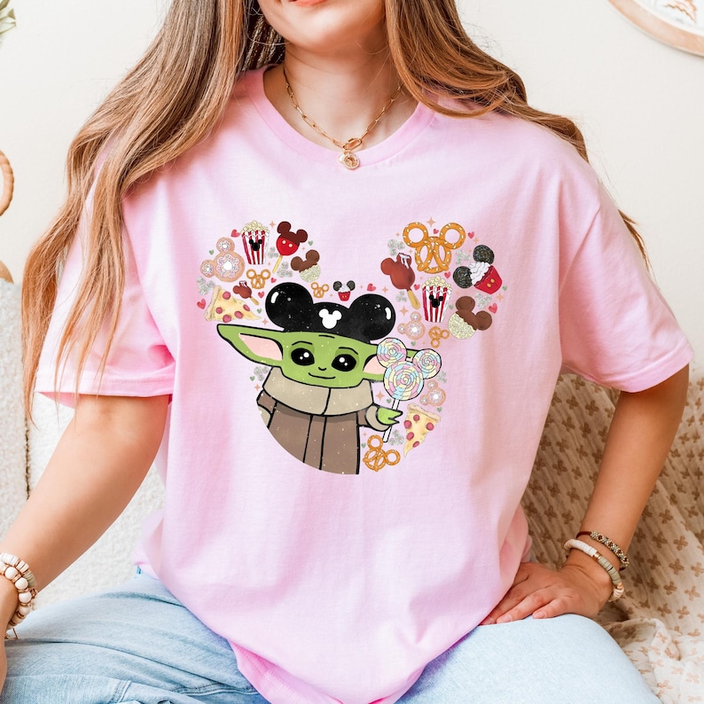 May include: A pale pink t-shirt with a graphic of Baby Yoda wearing Mickey Mouse ears and holding a lollipop. The design includes Mickey Mouse-themed snacks arranged in a heart shape.