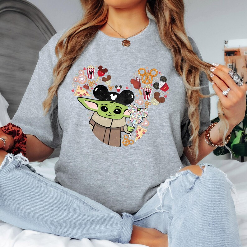 May include: A grey t-shirt with a graphic of Baby Yoda wearing Mickey Mouse ears and holding a lollipop. The design incorporates Mickey Mouse-themed treats such as popcorn, pretzels, and pizza, arranged in a heart shape.