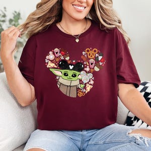 May include: Burgundy t-shirt with a Baby Yoda graphic wearing Mickey Mouse ears and holding a lollipop. The design is shaped like Mickey Mouse ears and filled with snacks. The shirt is made of a soft material.