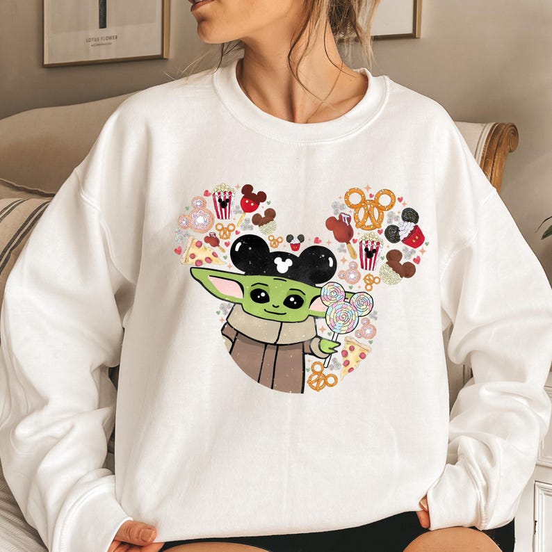 May include: White crew neck sweatshirt with a graphic of Grogu, or Baby Yoda, holding a lollipop. The design incorporates a Mickey Mouse head made of snacks. The sweatshirt is a casual style.