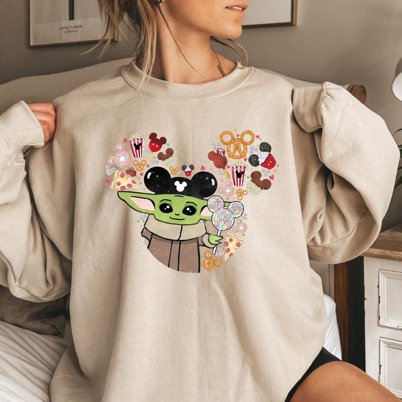 May include: Beige sweatshirt with a graphic of Baby Yoda wearing Mickey Mouse ears and holding a lollipop. The design includes a heart-shaped arrangement of Mickey Mouse-themed snacks and treats. The sweatshirt is a neutral colour.