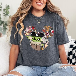 May include: Dark grey t-shirt with a graphic of Baby Yoda wearing Mickey Mouse ears and holding a lollipop. The design includes a Mickey Mouse head shape filled with snacks. The shirt is a comfortable, casual style.