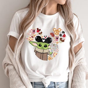 May include: White t-shirt with a cartoon alien character wearing Mickey Mouse ears and holding a lollipop. The design includes Mickey Mouse-themed food items such as popcorn, pretzels, and pizza, arranged in a heart shape.