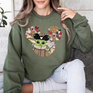 May include: Olive green sweatshirt with a Baby Yoda graphic wearing Mickey Mouse ears and holding a Mickey Mouse lollipop. The design incorporates Mickey Mouse-shaped treats and snacks. A fun, whimsical design.
