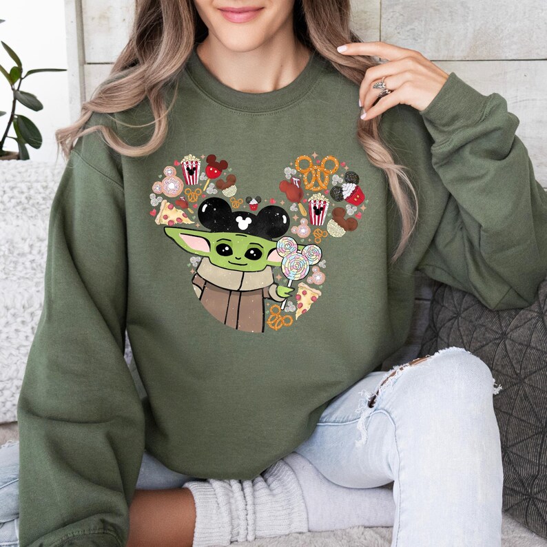 May include: Olive green sweatshirt with a Baby Yoda graphic wearing Mickey Mouse ears and holding a Mickey Mouse lollipop. The design incorporates Mickey Mouse-shaped treats and snacks. A fun, whimsical design.