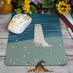 May include: A whimsical mousepad featuring a cartoon ghost in a field, with a white and gold wireless mouse. The mousepad has a teal and blue background with yellow accents. A pen and books are also visible.